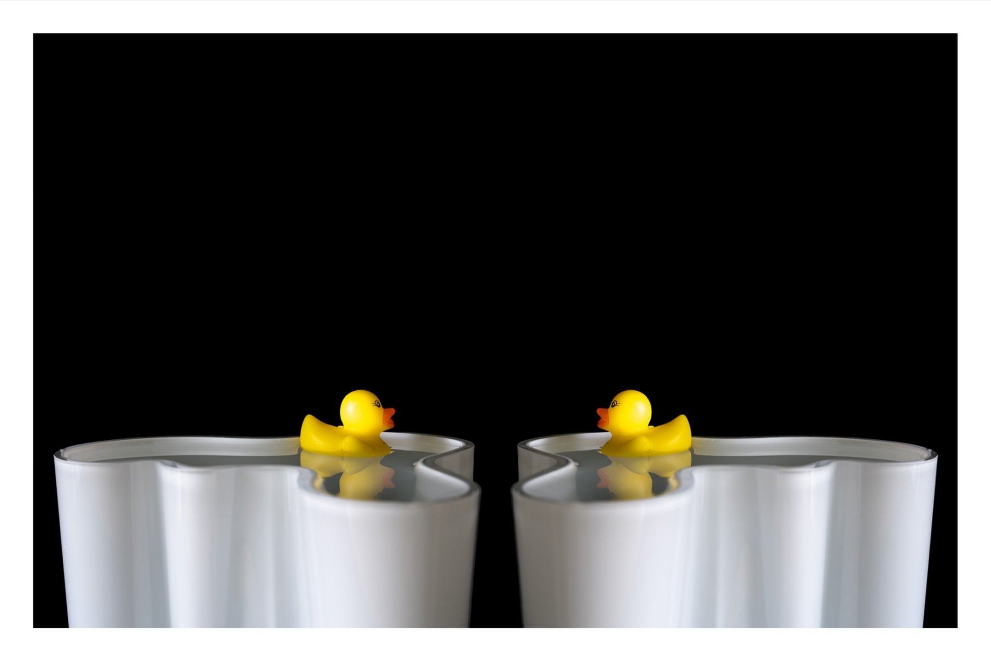 Two toy rubber ducks floating in an Alvar Aalto vase, facing each other in a soft, contemplative composition.