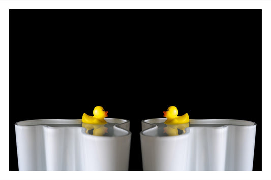 Two toy rubber ducks floating in an Alvar Aalto vase, facing each other in a soft, contemplative composition.