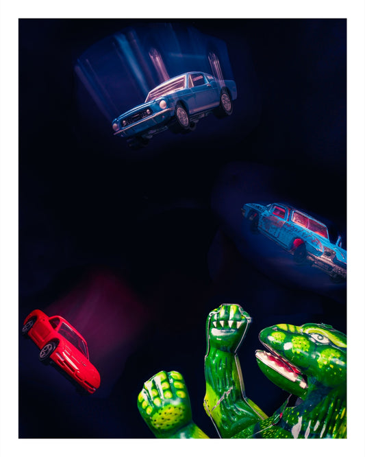 Toy Godzilla juggling Matchbox cars, including a red Corvette and a blue Mustang, in a dramatic and playful scene.
