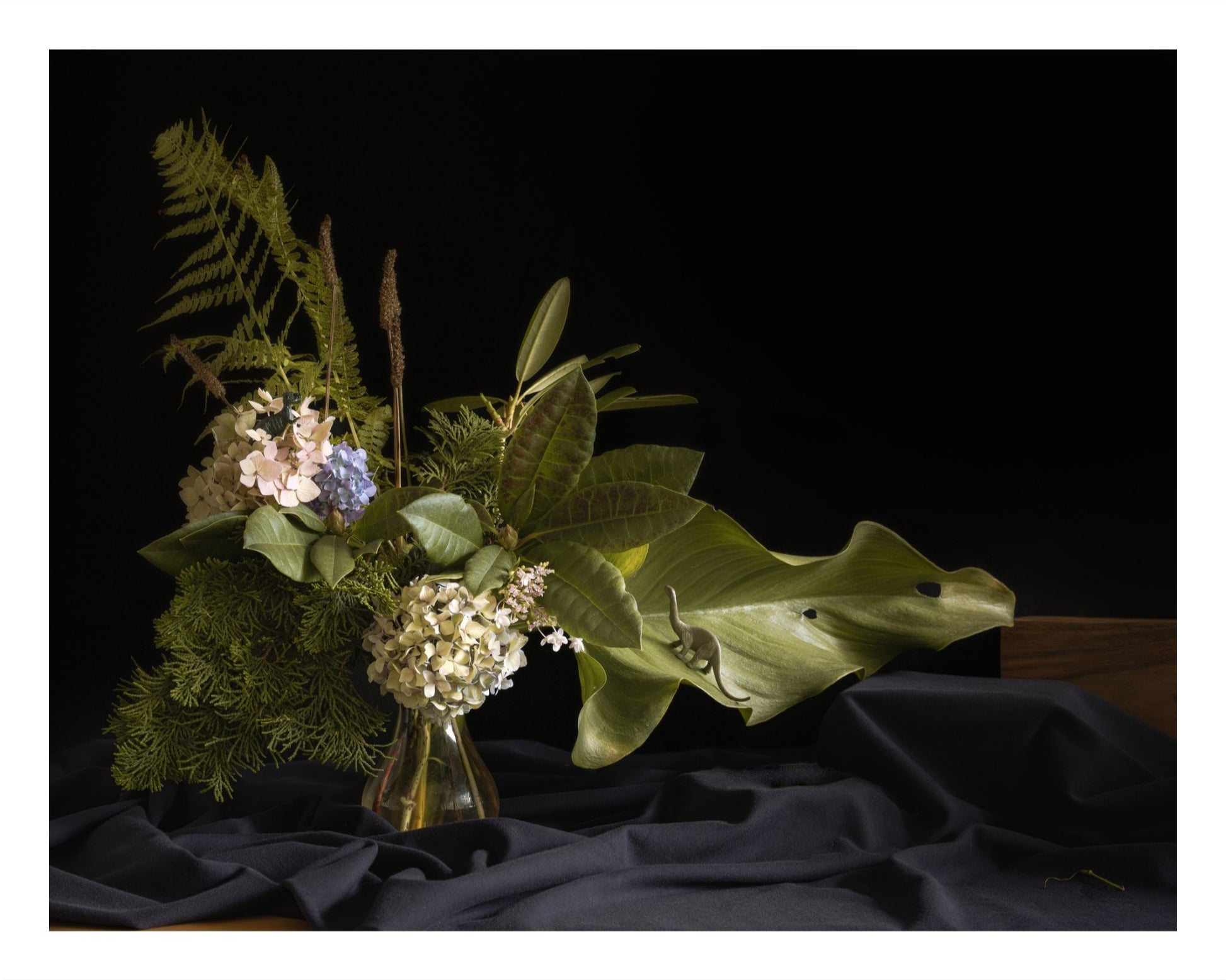 Color photograph of a floral arrangement with a brontosaurus and T. rex toy nestled among the flowers and foliage.
